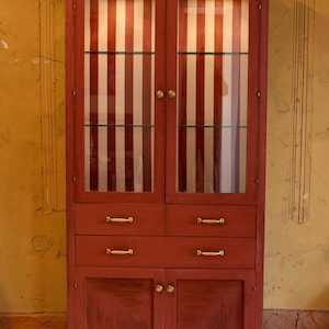 Op de afbeelding: Een rode houten kast met glazen deuren en messing hardware. De kastdeuren hebben een rood-wit gestreept patroon. De kast heeft twee glazen planken, twee laden en twee onderste deuren.