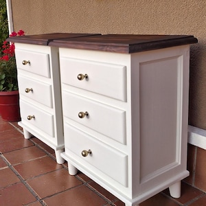 Puede incluir: Dos mesitas de noche blancas con tres cajones cada una, con tapas de madera marrón oscuro y pomos dorados. Las mesas tienen un diseño clásico con patas redondeadas. La altura es de aproximadamente 70 cm.