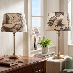 Op de afbeelding: Twee tafellampen met messing voetstukken en patronen. Eén kap heeft een vogelontwerp, de andere een vlinder. De lampen staan op een houten bureau met een notitieboek en pen.
