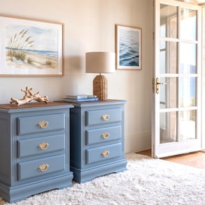 Peut inclure: Deux tables de chevet bleues à trois tiroirs avec des poignées dorées. Les dessus sont marron foncé. Elles sont posées sur un tapis blanc. Des œuvres d'art et une lampe sont visibles, avec vue sur la mer par une porte ouverte. Les tables mesurent environ 60 cm de large.