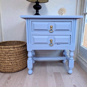 Puede incluir: Una mesita de noche pintada de azul claro con dos cajones y tiradores dorados. Una cesta tejida está a la izquierda. Una lámpara negra y una decoración de coral blanco están encima.