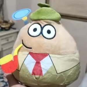 May include: A plush toy shaped like a tan, egg-shaped character wearing a green hat, a tan suit, and a red tie. It has large black eyes, a black smile, and a yellow and red pipe. A blue magnifying glass is attached to the hat.