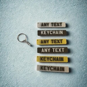 May include: A silver metal keyring next to six rectangular acrylic tags in white, yellow, and dark brown. The tags feature the text ANY TEXT or KEYCHAIN in dark lettering. Each tag has a small hole at one end for attachment to the keyring.