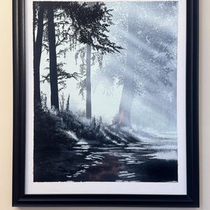 Op de afbeelding: Een ingelijste aquarel van een mistige bosscène. Het kunstwerk toont hoge bomen, een reflecterend water en lichtstralen die door de mist schijnen. De schildering is in grijstinten en zwart, met een zwarte lijst.