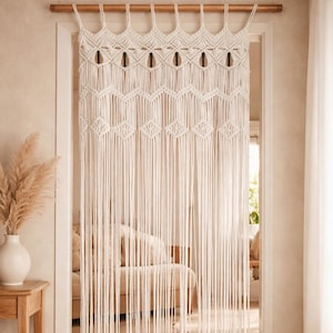 May include: Cream-colored macrame door curtain. The curtain is made of woven cotton with a geometric design. The curtain hangs from a wooden rod and is installed in a doorway. The curtain is a decorative element for a home.