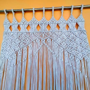 May include: A light blue macrame curtain with a geometric diamond pattern and fringe. The curtain is suspended from a silver rod, creating a decorative and textured window treatment.