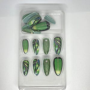 以下が含まれることがあります： 透明なプラスチック容器に入った、緑とグリッターのつけ爪のセット。爪には、グリッター、抽象的な模様、ラインストーンのアクセントなど、さまざまなデザインが施されています。爪はさまざまな緑の色合いです。