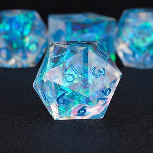 Puede incluir: Un conjunto de dados iridiscentes con numeración azul. Los dados poliédricos son transparentes con detalles internos azules y turquesas. Los dados están colocados sobre un fondo oscuro, con un dado enfocado y otros borrosos en el fondo.