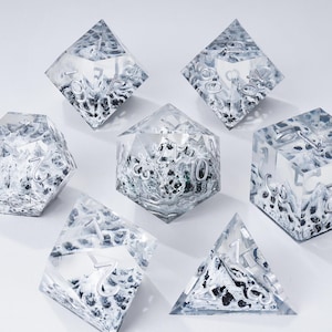 Peut inclure: Un ensemble de sept d&eacute;s en r&eacute;sine transparente avec des chiffres argent&eacute;s et un motif int&eacute;rieur textur&eacute; noir et blanc. Les d&eacute;s sont de formes g&eacute;om&eacute;triques vari&eacute;es, dont un d4, d6, d8, d10, d12, d20 et un d100.