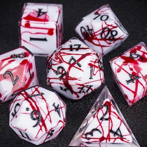 May include: A collection of seven polyhedral dice. Each die has a white base with a red splatter pattern and black numbers. The set includes a D4, D6, D8, D10, D12, D20, and a percentile die. Ideal for role-playing games.