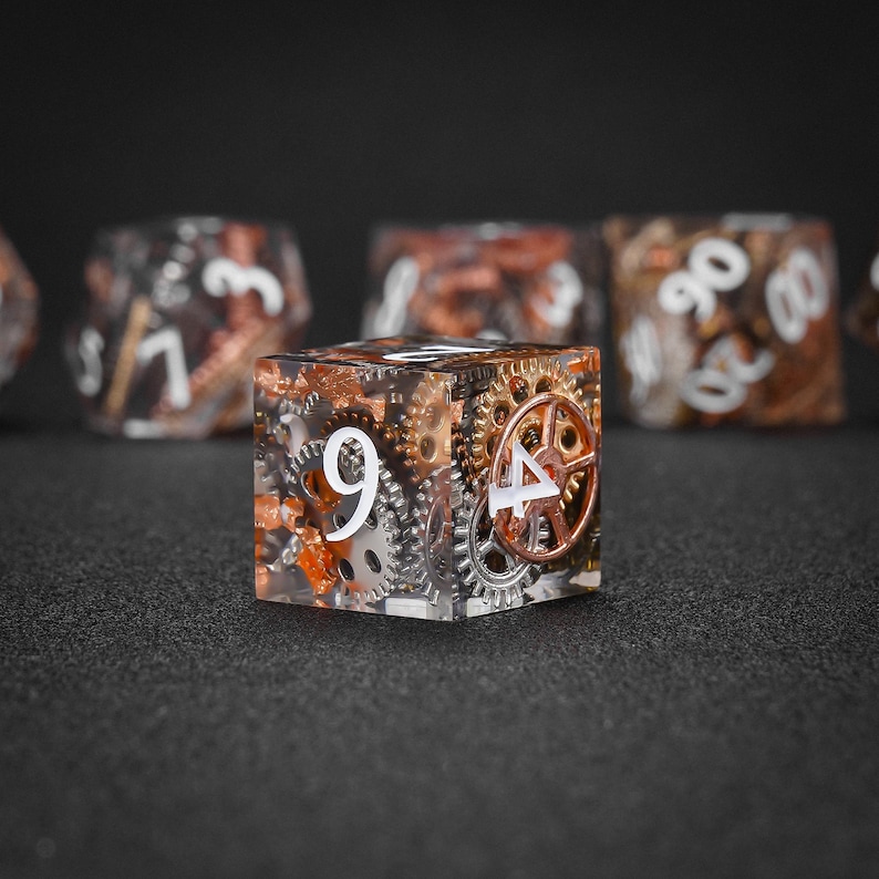 May include: A clear resin die with embedded gears and metallic accents. The die features the number 9 and 4 in white, with other dice visible in the background. The dice have a steampunk aesthetic.