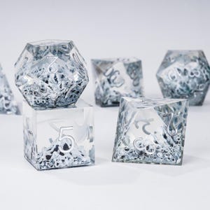 Peut inclure: Ensemble de d&eacute;s transparents &agrave; multiples faces, avec des chiffres argent&eacute;s et un int&eacute;rieur textur&eacute; et neigeux. Ces d&eacute;s, con&ccedil;us pour les jeux de soci&eacute;t&eacute;, sont pr&eacute;sent&eacute;s sur une surface blanche. L'ensemble comprend diff&eacute;rentes formes, comme un cube et un d&eacute; &agrave; 20 faces.