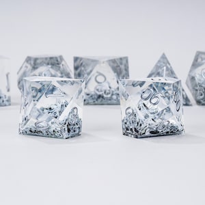 Peut inclure: Un ensemble de d&eacute;s transparents &agrave; multiples faces, avec des chiffres argent&eacute;s et un int&eacute;rieur sombre et textur&eacute;. Les d&eacute;s sont dispos&eacute;s sur une surface blanche, mettant en valeur leurs formes g&eacute;om&eacute;triques et leurs d&eacute;tails complexes. Pour les jeux de soci&eacute;t&eacute;.