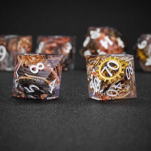 May include: Close-up of several steampunk-themed dice. The dice are clear with embedded gears, cogs, and other metallic components in shades of gold, copper, and silver. White numbers are visible on the faces. The dice are set against a dark background.