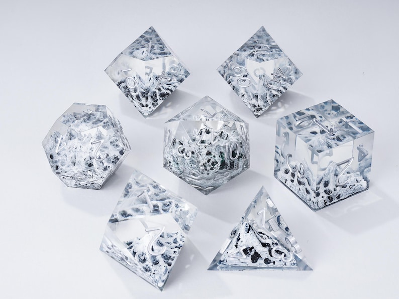 Peut inclure: Un ensemble de sept d&eacute;s en r&eacute;sine transparente avec des chiffres argent&eacute;s et un motif int&eacute;rieur textur&eacute; noir et blanc. Les d&eacute;s sont de formes g&eacute;om&eacute;triques vari&eacute;es, dont un d4, d6, d8, d10, d12, d20 et un d100.