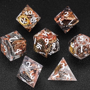 May include: Set of seven polyhedral dice with a steampunk theme. The dice are clear with embedded gears in copper, gold, and silver. White numbers are visible on each face. The dice are arranged on a black surface.