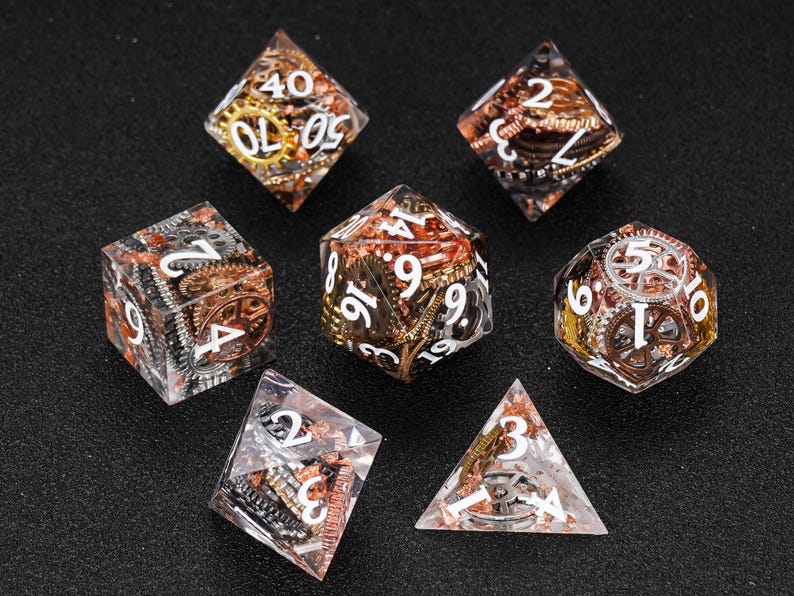May include: Set of seven polyhedral dice with a steampunk theme. The dice are clear with embedded gears in copper, gold, and silver. White numbers are visible on each face. The dice are arranged on a black surface.