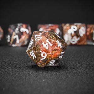 May include: A clear, multi-sided die with white numbers and a steampunk design. The die features gears and copper-colored accents. Other dice are visible in the background, all with a similar design. The dice are for tabletop games.