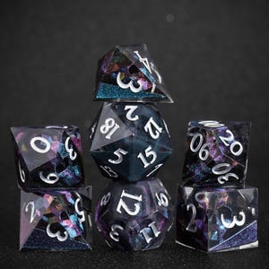 Peut inclure: Un ensemble de sept d&eacute;s poly&eacute;driques de diff&eacute;rentes formes. Ils sont faits d'un mat&eacute;riau sombre et translucide avec un m&eacute;lange tourbillonnant de paillettes violettes, bleues et dor&eacute;es. Les chiffres sont peints en blanc.