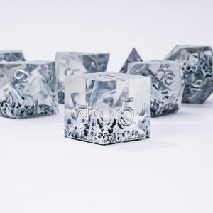 Peut inclure: Ensemble de d&eacute;s en r&eacute;sine transparente avec des chiffres argent&eacute;s et un int&eacute;rieur textur&eacute; et glac&eacute;. Les d&eacute;s sont de formes vari&eacute;es, dont un cube, une pyramide et un d&eacute; &agrave; plusieurs faces. Les d&eacute;s sont pos&eacute;s sur un fond blanc.