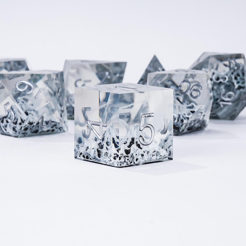 Peut inclure: Ensemble de d&eacute;s en r&eacute;sine transparente avec des chiffres argent&eacute;s et un int&eacute;rieur textur&eacute; et glac&eacute;. Les d&eacute;s sont de formes vari&eacute;es, dont un cube, une pyramide et un d&eacute; &agrave; plusieurs faces. Les d&eacute;s sont pos&eacute;s sur un fond blanc.