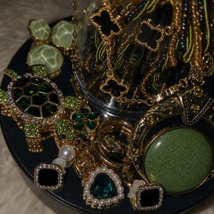 Op de afbeelding: Een assortiment sieraden, waaronder kettingen, oorbellen, ringen en hangers. De stukken hebben gouden tinten, groene edelstenen en zwarte accenten. Een decoratieve hanger in de vorm van een schildpad en klaver is zichtbaar.