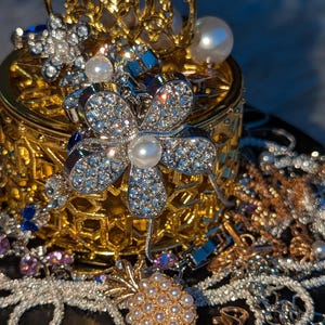 May include: A gold-toned metal container with an intricate lattice design, topped with a decorative filigree element. The container is surrounded by various jewellery pieces, including a large flower-shaped brooch with clear crystals and a central pearl.