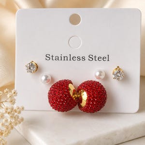 May include: A set of stainless steel earrings on a white display card featuring two large spherical studs covered in red crystals, two small white faux pearl studs, and two clear round crystal studs with gold-toned metal settings.