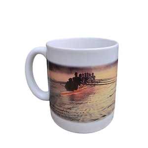 Op de afbeelding: Een witte keramische mok met een fotoprint van een roeiteam in een lange boot op het water bij zonsondergang. De afbeelding toont meerdere roeiers met uitgestrekte riemen, wat een silhouet vormt tegen het warme, gouden licht op het water.
