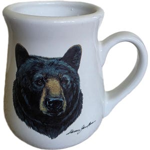 以下が含まれることがあります： 黒いクマの顔のイラストが描かれた白いセラミックマグカップ。 クマは茶色の鼻をしており、白い背景に描かれています。 マグカップにはカーブしたハンドルと、底にサインがあります。 自然愛好家への素晴らしい贈り物です。