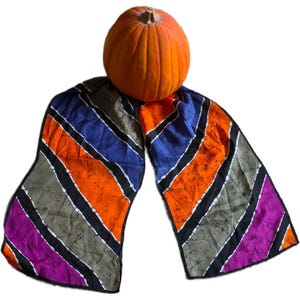 May include: A rectangular scarf featuring a diagonal striped pattern in vibrant shades of orange, blue, gray, and purple. Each stripe is separated by a thin black border with white accents. A round, orange pumpkin rests on top of the fabric.