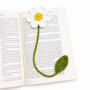 Könnte beinhalten: Ein handgefertigtes Häkel-Lesezeichen in Form einer weißen Blume mit leuchtend gelber Mitte, befestigt an einem langen grünen Stiel, der in einem einzelnen grünen Blatt endet. Das Lesezeichen liegt in einem aufgeschlagenen Buch mit Text.