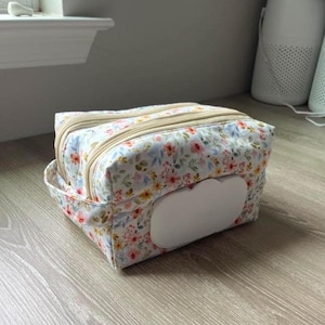 May include: Floral print diaper wipe case with a white wipe dispenser. The case has a beige zipper and a floral pattern with pink, yellow, and blue flowers on a white background. The case is rectangular with a curved edge.