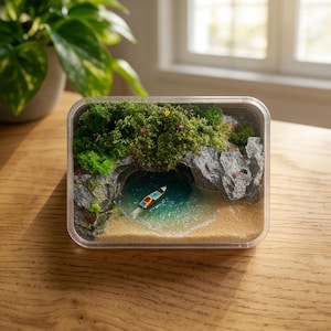 Könnte beinhalten: Miniatur-Diorama einer tropischen Strandszene in einem transparenten rechteckigen Behälter. Die Szene umfasst ein kleines Boot, einen Sandstrand, türkisfarbenes Wasser, Felsklippen und üppige grüne Vegetation. Das Diorama steht auf einem Holztisch.