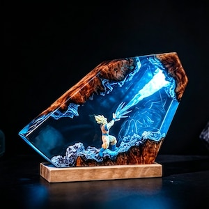 May include: A resin sculpture featuring a character in a fighting pose, emitting a blue energy blast. The sculpture is encased in a clear, blue-tinted resin with a wooden base and brown, textured edges. The artwork is illuminated with blue light.