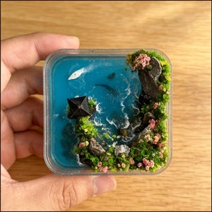 May include: Miniature diorama scene in a clear square container. The scene depicts a blue ocean with a small black structure, a white boat, and a rocky island with green moss and pink flowers. The diorama is held in a hand.