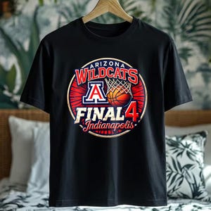 May include: Black t-shirt featuring a circular graphic with the text "ARIZONA WILDCATS FINAL 4 Indianapolis". The graphic includes a basketball, net, and the letter "A" in a red, white, and blue design. The shirt is hanging on a wooden hanger.