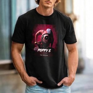 Puede incluir: Camiseta negra con un gráfico de un personaje de Poppy Playtime. El diseño incluye una figura de bufón roja y blanca y el texto "POPPY 5 PLAYTIME THE PROTOTYPE".