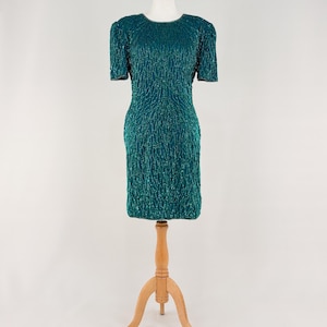 Peut inclure: Robe à paillettes turquoise à manches courtes et col rond. La robe est présentée sur un mannequin en bois. La robe présente un motif vertical texturé et une silhouette ajustée.