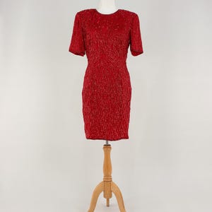 Peut inclure: Robe rouge à perles, à manches courtes et col rond. La robe est présentée sur un mannequin en bois. Silhouette ajustée et surface texturée, idéale pour une soirée ou un événement spécial.