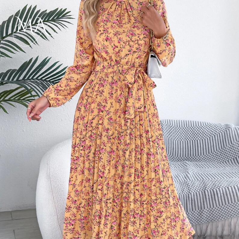Puede incluir: Vestido amarillo de manga larga con estampado floral en rosa y morado. El vestido presenta una falda plisada, una cintura anudada y un cuello alto. El vestido es de largo midi.