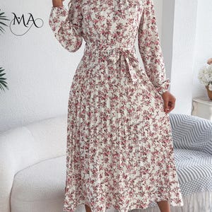 Puede incluir: Vestido marfil con estampado floral rosa, mangas largas y falda plisada. Tiene cintura anudada y cuello alto. Las letras "MA" son visibles en la esquina superior izquierda.