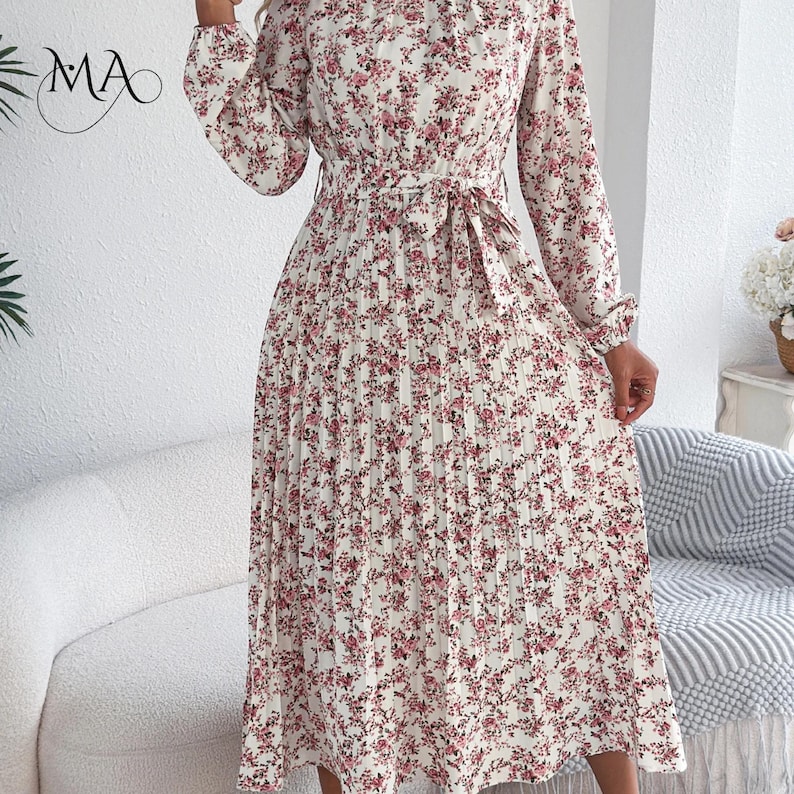 Puede incluir: Vestido marfil con estampado floral rosa, mangas largas y falda plisada. Tiene cintura anudada y cuello alto. Las letras "MA" son visibles en la esquina superior izquierda.
