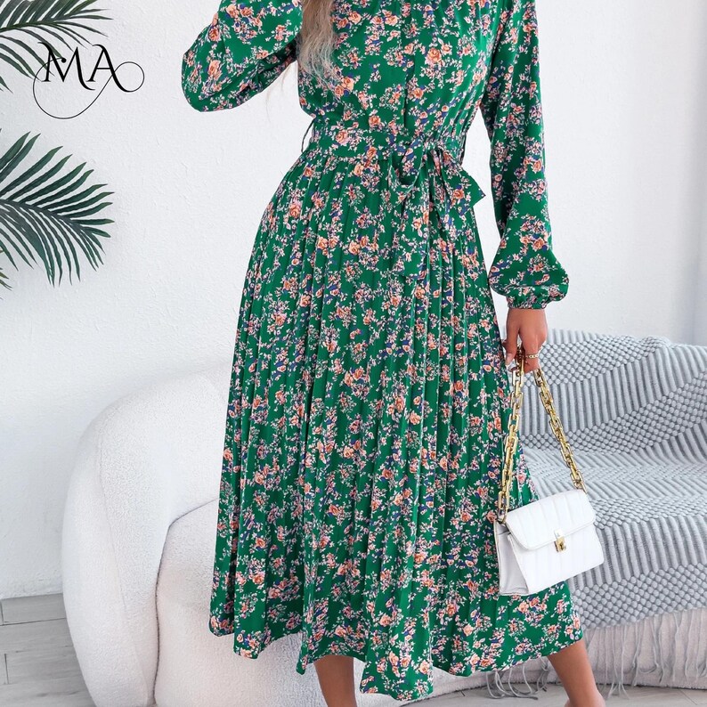 Puede incluir: Vestido midi verde con estampado floral, mangas largas y falda plisada. El vestido tiene una cintura anudada y un cuello alto. Se complementa con un bolso blanco acolchado con cadena dorada. El vestido mide aproximadamente 110 cm de largo.