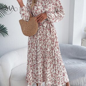 Puede incluir: Un vestido plisado de manga larga con un estampado floral en tonos rosas y rojos sobre fondo blanco. El vestido tiene una cintura anudada y se combina con un bolso de mano tejido. El vestido tiene un cuello alto.