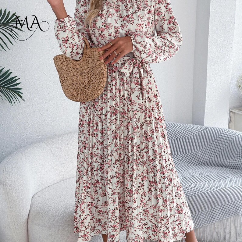 Puede incluir: Un vestido plisado de manga larga con un estampado floral en tonos rosas y rojos sobre fondo blanco. El vestido tiene una cintura anudada y se combina con un bolso de mano tejido. El vestido tiene un cuello alto.