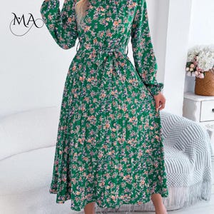 Puede incluir: Un vestido verde con estampado floral, mangas largas y cintur&oacute;n anudado. El vestido presenta un cuello alto y una falda plisada. El estampado floral incluye flores rosas, naranjas y azules sobre un fondo verde. El vestido es hasta la rodilla y parece ser de tejido ligero.