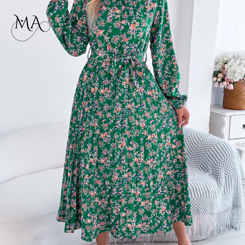 Puede incluir: Un vestido verde con estampado floral, mangas largas y cintur&oacute;n anudado. El vestido presenta un cuello alto y una falda plisada. El estampado floral incluye flores rosas, naranjas y azules sobre un fondo verde. El vestido es hasta la rodilla y parece ser de tejido ligero.