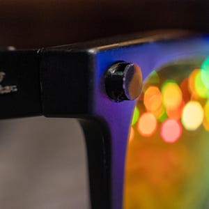 May include: Close-up of black sunglasses with the Ray-Ban logo on the temple. The lenses reflect colorful, blurred lights. The frame is black with a small silver rivet.