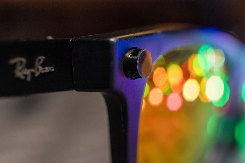 May include: Close-up of black sunglasses with the Ray-Ban logo on the temple. The lenses reflect colorful, blurred lights. The frame is black with a small silver rivet.